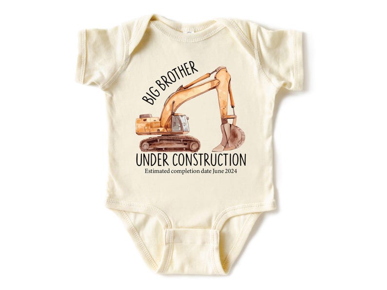 Pode incluir: Body de beb&eacute; creme com uma ilustra&ccedil;&atilde;o em aquarela de uma escavadeira. O texto diz "IRM&Atilde;O MAIOR EM CONSTRU&Ccedil;&Atilde;O" com uma data de conclus&atilde;o estimada em junho de 2024.