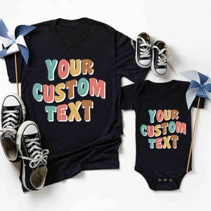 May include: Two black t-shirts with the text "YOUR CUSTOM TEXT" in colorful, outlined letters. The larger shirt is a short-sleeved t-shirt, and the smaller shirt is a bodysuit.