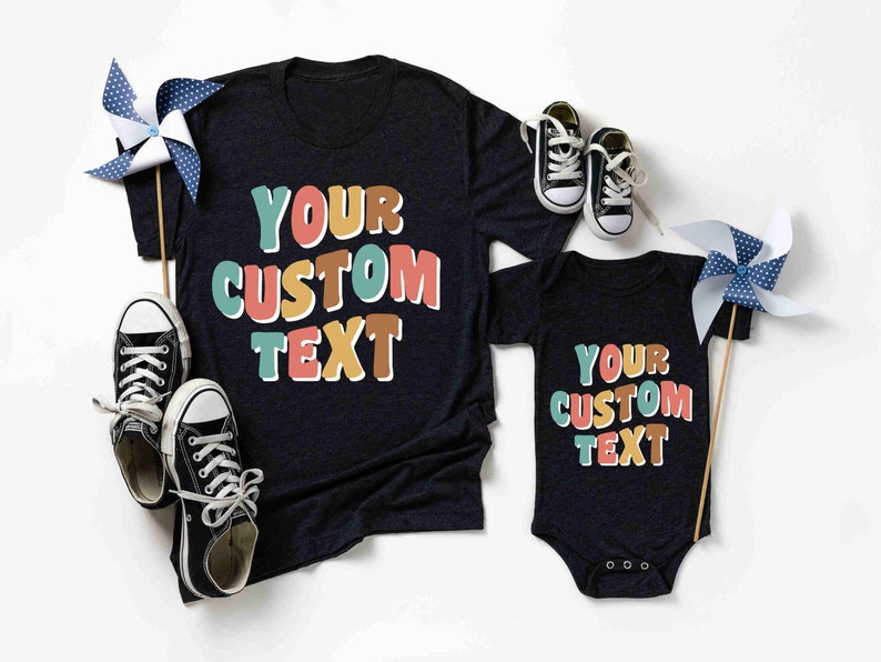 May include: Two black t-shirts with the text "YOUR CUSTOM TEXT" in colorful, outlined letters. The larger shirt is a short-sleeved t-shirt, and the smaller shirt is a bodysuit.