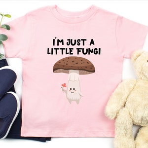May include: Pink toddler t-shirt with the text "I'M JUST A LITTLE FUNGI" above a cartoon mushroom character holding a flower. The mushroom has a brown cap and a white stem. Dark blue pants and shoes are next to the shirt.