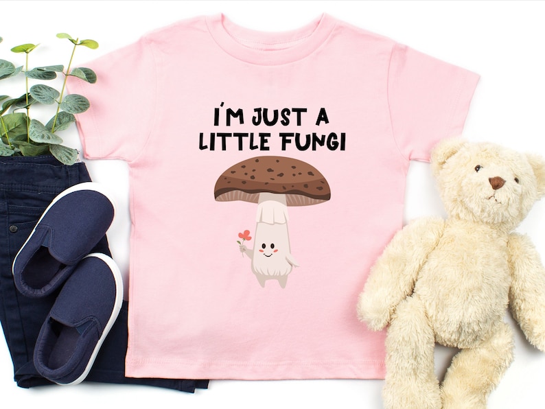 May include: Pink toddler t-shirt with the text "I'M JUST A LITTLE FUNGI" above a cartoon mushroom character holding a flower. The mushroom has a brown cap and a white stem. Dark blue pants and shoes are next to the shirt.