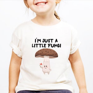 May include: Off-white t-shirt featuring the text "I'M JUST A LITTLE FUNGI" above a cartoon mushroom holding a flower. The mushroom has a brown cap with white spots and a light beige stem. The shirt is on a child.
