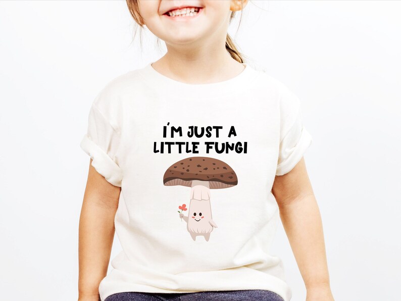 May include: Off-white t-shirt featuring the text "I'M JUST A LITTLE FUNGI" above a cartoon mushroom holding a flower. The mushroom has a brown cap with white spots and a light beige stem. The shirt is on a child.