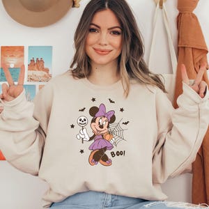 K&ouml;nnte beinhalten: Ein beigefarbenes Sweatshirt mit Rundhalsausschnitt und einem Aufdruck von Minnie Maus in einem lila Kleid mit passender Schleife, die einen Totenkopf-Lutscher und einen K&uuml;rbis h&auml;lt. Das Motiv zeigt zudem ein Spinnennetz, Flederm&auml;use, Sterne und den Schriftzug BOO.
