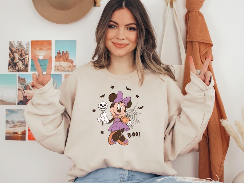 K&ouml;nnte beinhalten: Ein beigefarbenes Sweatshirt mit Rundhalsausschnitt und einem Aufdruck von Minnie Maus in einem lila Kleid mit passender Schleife, die einen Totenkopf-Lutscher und einen K&uuml;rbis h&auml;lt. Das Motiv zeigt zudem ein Spinnennetz, Flederm&auml;use, Sterne und den Schriftzug BOO.