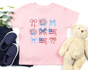 May include: A light pink toddler t-shirt featuring watercolor illustrations of bows, fireworks, and American flags in red, white, and blue. The shirt is paired with navy blue pants and shoes, and a teddy bear.