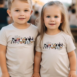 K&ouml;nnte beinhalten: Zwei Kinder tragen beige T-Shirts mit einem schwarz-wei&szlig;en Motiv von Mickey Mouse und seinen Freunden in Halloween-Kost&uuml;men. Das Motiv zeigt Mickey Mouse, Minnie Mouse, Donald Duck, Goofy, Pluto und eine Hexe.