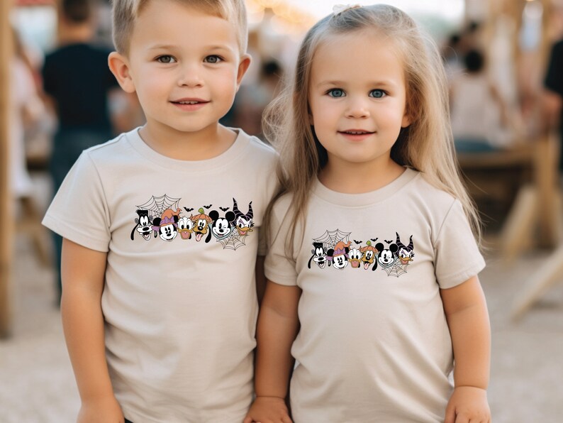 K&ouml;nnte beinhalten: Zwei Kinder tragen beige T-Shirts mit einem schwarz-wei&szlig;en Motiv von Mickey Mouse und seinen Freunden in Halloween-Kost&uuml;men. Das Motiv zeigt Mickey Mouse, Minnie Mouse, Donald Duck, Goofy, Pluto und eine Hexe.