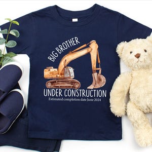 Pode incluir: Camiseta azul marinho para crian&ccedil;as com uma ilustra&ccedil;&atilde;o em aquarela de uma escavadeira de constru&ccedil;&atilde;o. A camisa diz "Irm&atilde;o Mais Velho em Constru&ccedil;&atilde;o" com uma data de conclus&atilde;o estimada em junho de 2024. Inclui sapatos, cal&ccedil;as e um ursinho de pel&uacute;cia azul marinho.