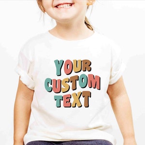 May include: White t-shirt with the text "YOUR CUSTOM TEXT" in colorful, outlined letters. The letters are in shades of teal, pink, orange, and brown.