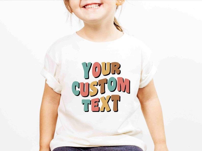May include: White t-shirt with the text "YOUR CUSTOM TEXT" in colorful, outlined letters. The letters are in shades of teal, pink, orange, and brown.