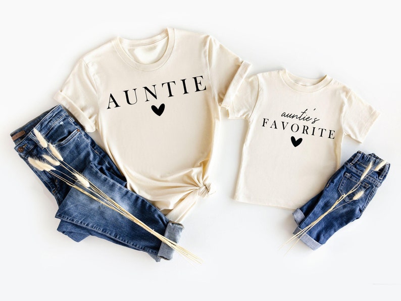 May include: Two cream coloured t-shirts with black text. The larger shirt says "AUNTIE" with a black heart. The smaller shirt says "auntie's FAVORITE" with a black heart.
