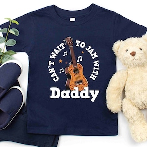 May include: A navy blue t-shirt with a white graphic of a ukulele and the text "Can't Wait To Jam With Him Daddy".