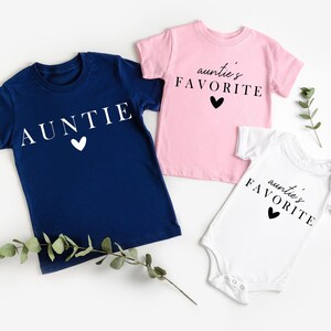 May include: Three t-shirts in different colours and designs. A navy blue t-shirt with the word "AUNTIE" in white text and a white heart. A pink t-shirt with the text "auntie's FAVORITE" in black text and a black heart. A white t-shirt with the text "auntie's FAVORITE" in black text and a black heart.