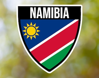 Könnte beinhalten: Ein weißer Aufkleber mit schwarzem Rand und dem Wort "NAMIBIA" in weißen Buchstaben. Der Aufkleber zeigt die namibische Flagge, die ein blaues Feld mit einer gelben Sonne in der oberen Mastspitze und einem roten Diagonalstreifen von der unteren Mastspitze zur oberen Flugspitze hat, mit einem grünen Streifen unter dem roten Streifen.