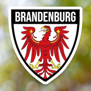 May include: A sticker featuring the Brandenburg coat of arms. The design includes a red eagle with yellow accents on a white background, framed by a black border. The word "BRANDENBURG" is displayed in white letters on a black banner.
