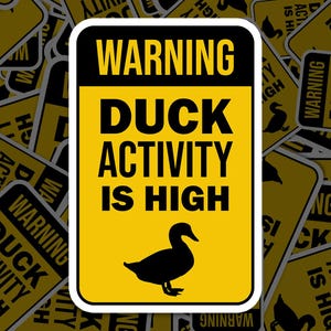 Op de afbeelding: Een gele sticker met een zwarte rand en de woorden "WARNING DUCK ACTIVITY IS HIGH" in vette zwarte letters. Een zwart silhouet van een eend staat onderaan. De sticker is omgeven door andere soortgelijke stickers.