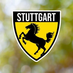 Pode incluir: Um adesivo em forma de escudo amarelo e preto com a palavra "STUTTGART" em letras brancas no topo e uma silhueta preta de um cavalo empinado dentro.