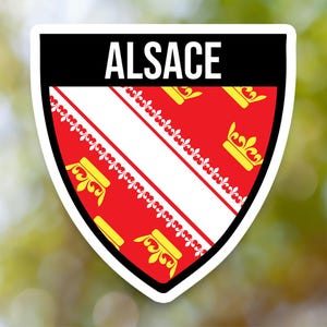 May include: A shield-shaped sticker featuring the word "ALSACE" in white on a black banner. The shield has a red background with a white stripe and yellow crowns, representing the Alsace region.