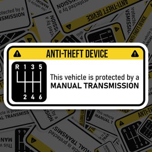 May include: A yellow and black warning sticker with a white exclamation point and the text "ANTI-THEFT DEVICE". The sticker also features a black and white illustration of a manual transmission gear shift pattern with the numbers 1, 2, 3, 4, 5, and 6. The text "This vehicle is protected by a MANUAL TRANSMISSION" is also included on the sticker.