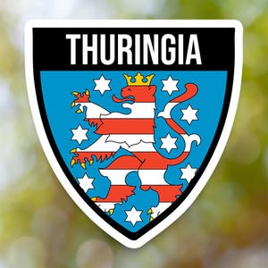 May include: A sticker featuring the word "THURINGIA" in white text on a black background. Below, a blue shield displays a red and white striped lion with a gold crown, surrounded by white stars.