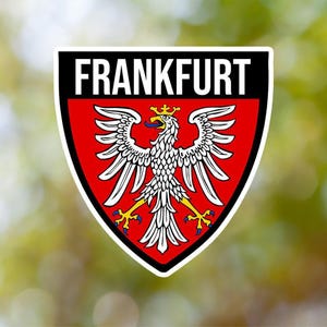 May include: A red and white shield with the word "FRANKFURT" in white letters on a black background. A white eagle with a gold crown and yellow beak is centred on the shield.