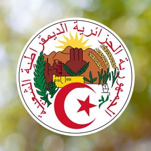Könnte beinhalten: Ein weißes rundes Siegel mit arabischem Text um den Rand. Die Mitte zeigt einen roten Halbmond mit einem weißen fünfzackigen Stern, ein gelbes Gebäude, grüne Bäume und eine braune Bergkette mit einer gelben Sonne.