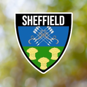 May include: A black and white shield-shaped sticker with the word "SHEFFIELD" in white letters at the top. The shield has a blue background with a silver design of arrows and a green background with three yellow sheaves of wheat.