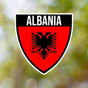 Puede incluir: Una pegatina en forma de escudo en blanco y negro con la palabra "ALBANIA" en letras blancas en la parte superior. El escudo tiene un fondo rojo con un águila bicéfala negra en el centro.