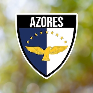 May include: A black and white shield-shaped sticker with the word "AZORES" in white letters at the top. The shield features a blue and white background with a yellow bird in the center and a ring of yellow stars around the blue section.