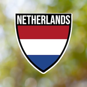 Könnte beinhalten: Ein schildförmiger Aufkleber mit dem Wort "NIEDERLANDE" in schwarzem Text oben. Das Schild ist in drei horizontale Streifen unterteilt: rot, weiß und blau.