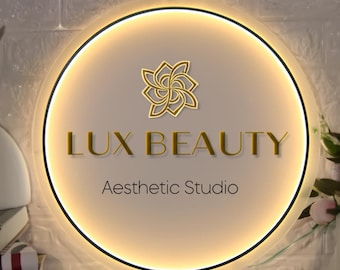 Puede incluir: Letrero circular iluminado con el texto "LUX BEAUTY" en letras doradas, encima de "Aesthetic Studio". El letrero presenta un diseño floral dorado y un borde negro, con un brillo cálido y acogedor.