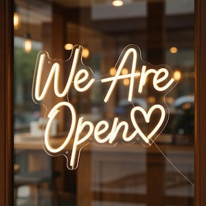 Op de afbeelding: Een neonbord in een etalage toont de woorden "We Are Open" in een cursief lettertype, met een hartje in plaats van de "o" in "Open". Het bord straalt een warme, uitnodigende gloed uit.