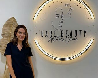 May include: A circular, illuminated wall sign featuring the text BARE BEAUTY Aesthetics Clinic in black lettering, accompanied by a minimalist line-art illustration of two faces and scattered dots, mounted on a wall with a warm, glowing light border.
