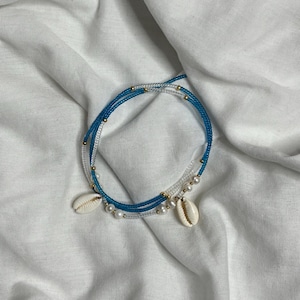 Peut inclure: Un bracelet de perles superposées avec un design bleu et transparent. Le bracelet comporte de petites perles dorées, des perles blanches et deux coquillages blancs. Le bracelet est présenté sur une surface blanche texturée.