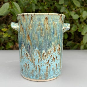 May include: A handmade ceramic vase with a cylindrical shape and two small handles. The vase features a turquoise and brown glaze with a dripped effect. The ceramic piece is suitable for home decor.