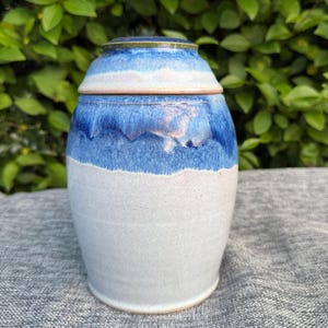 以下が含まれることがあります： 蓋付きの陶器の壺。2色のデザインが特徴です。上部は鮮やかな青色で、明るい青と白のベースに変わります。この壺はおそらく手作りで、ユニークで芸術的な美学を示しています。