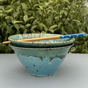 May include: Two teal blue ceramic bowls with a speckled glaze and a wooden chopstick resting on top. The bowls are stacked on top of each other.
