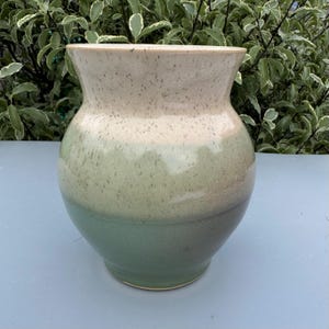 Può includere: Un vaso in ceramica fatto a mano con un design bicolore maculato. La parte superiore è beige chiaro, che sfuma in un verde tenue al centro e in un verde più scuro alla base. Il vaso ha un'ampia bocca e un corpo arrotondato.