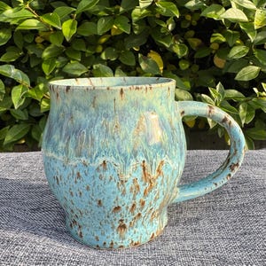 May include: A handcrafted ceramic mug with a rounded body and a large handle. The mug has a turquoise and brown glaze with a speckled pattern. The mug is suitable for hot or cold drinks.