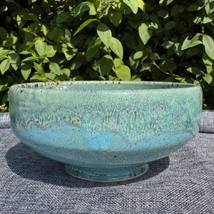 Peut inclure: Un bol en céramique émaillée turquoise. Le bol a une forme large et peu profonde avec une base à pied. L'émail a un aspect texturé et moucheté, avec des variations de couleur du bleu clair au turquoise foncé. Idéal pour servir ou décorer. Diamètre: environ 25 cm.