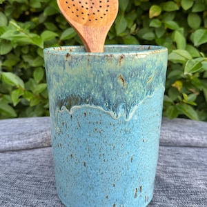 May include: A turquoise and blue ceramic utensil holder with a wooden slotted spoon. The holder has a textured surface and a glossy finish. Ideal for kitchen storage, the holder is approximately 15 cm tall.