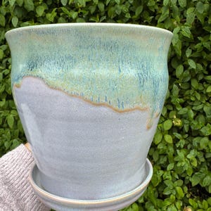 May include: A ceramic plant pot with a matching saucer. The pot is light blue with a turquoise and gold glaze on the top half. It has a wide opening and a rounded shape. The pot is suitable for indoor or outdoor use.
