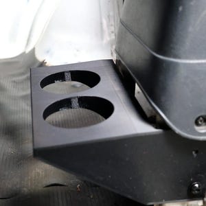 May include: A black plastic dual cup holder attachment designed for a vehicle center console. The unit features two circular cutouts for beverage containers and is secured with visible screws to the side of a dark gray automotive console component.
