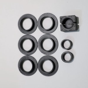 Könnte beinhalten: Eine Sammlung dunkelgrauer 3D-gedruckter Komponenten, bestehend aus sechs kreisförmigen Ringen mit breiten flachen Rändern, zwei kleineren einfachen Ringen und einem quadratischen Block mit kreisförmigem Ausschnitt und zentraler Vertiefung.