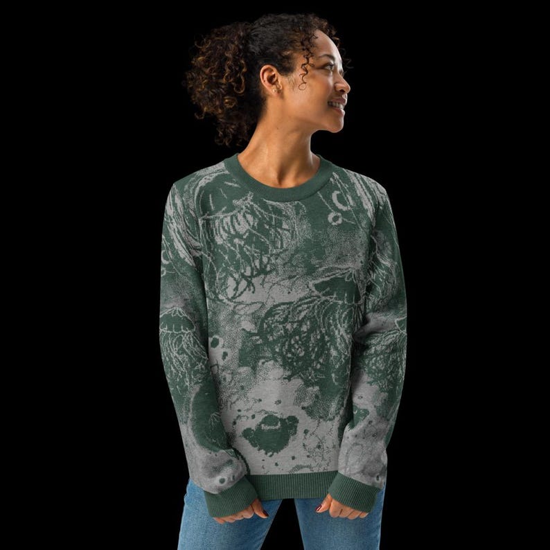 K&ouml;nnte beinhalten: Ein gr&uuml;nes und graues Sweatshirt mit einem Quallen-Design. Der lang&auml;rmlige Pullover hat einen Rundhalsausschnitt und gerippte B&uuml;ndchen und Saum. Das Quallenmuster ist eine Mischung aus Dunkelgr&uuml;n und Grau und erzeugt einen strukturierten, k&uuml;nstlerischen Look. Der Hintergrund ist schwarz.