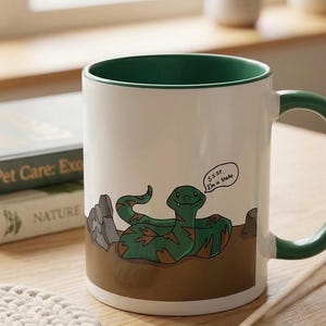 Puede incluir: Una taza de cerámica blanca con interior y asa verdes, que presenta una ilustración de dibujos animados de una serpiente verde con manchas marrones sobre un suelo marrón. La serpiente tiene un bocadillo que dice "SS-SP... I'm a snake."