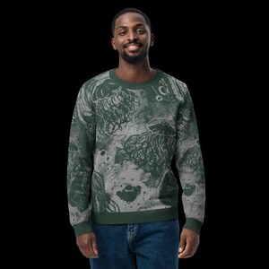 K&ouml;nnte beinhalten: Ein gr&uuml;nes und graues Sweatshirt mit Rundhalsausschnitt und einem Quallenmuster. Das Design hat einen verwitterten, k&uuml;nstlerischen Look. Das Sweatshirt wird von einer Person mit dunklen Haaren und Bart getragen, die blaue Jeans tr&auml;gt.