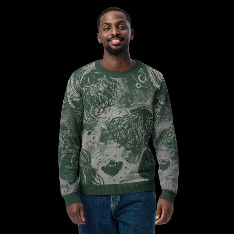K&ouml;nnte beinhalten: Ein gr&uuml;nes und graues Sweatshirt mit Rundhalsausschnitt und einem Quallenmuster. Das Design hat einen verwitterten, k&uuml;nstlerischen Look. Das Sweatshirt wird von einer Person mit dunklen Haaren und Bart getragen, die blaue Jeans tr&auml;gt.