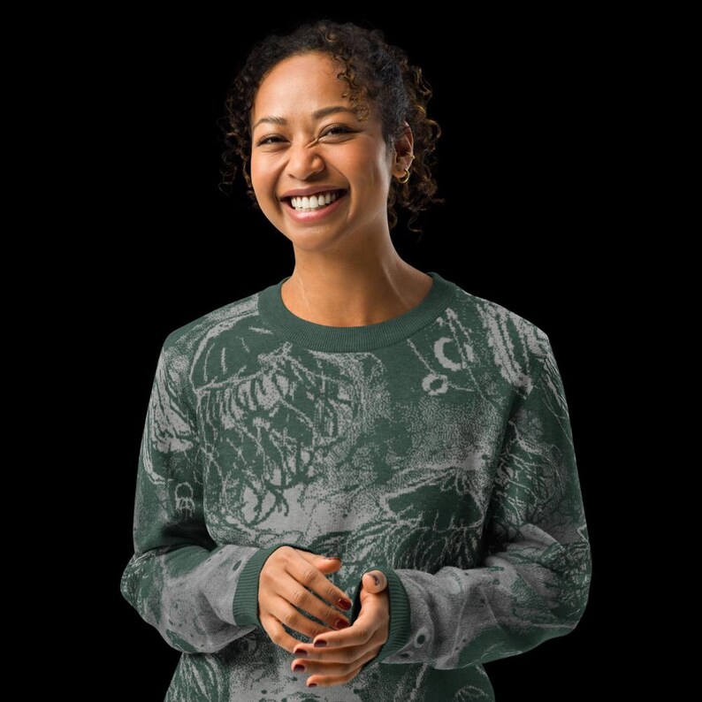 K&ouml;nnte beinhalten: Ein gr&uuml;nes Sweatshirt mit Rundhalsausschnitt und einem abstrakten wei&szlig;en und grauen Design. Das Sweatshirt hat lange &Auml;rmel und einen gerippten Kragen und B&uuml;ndchen. Das Model l&auml;chelt und tr&auml;gt das Sweatshirt.